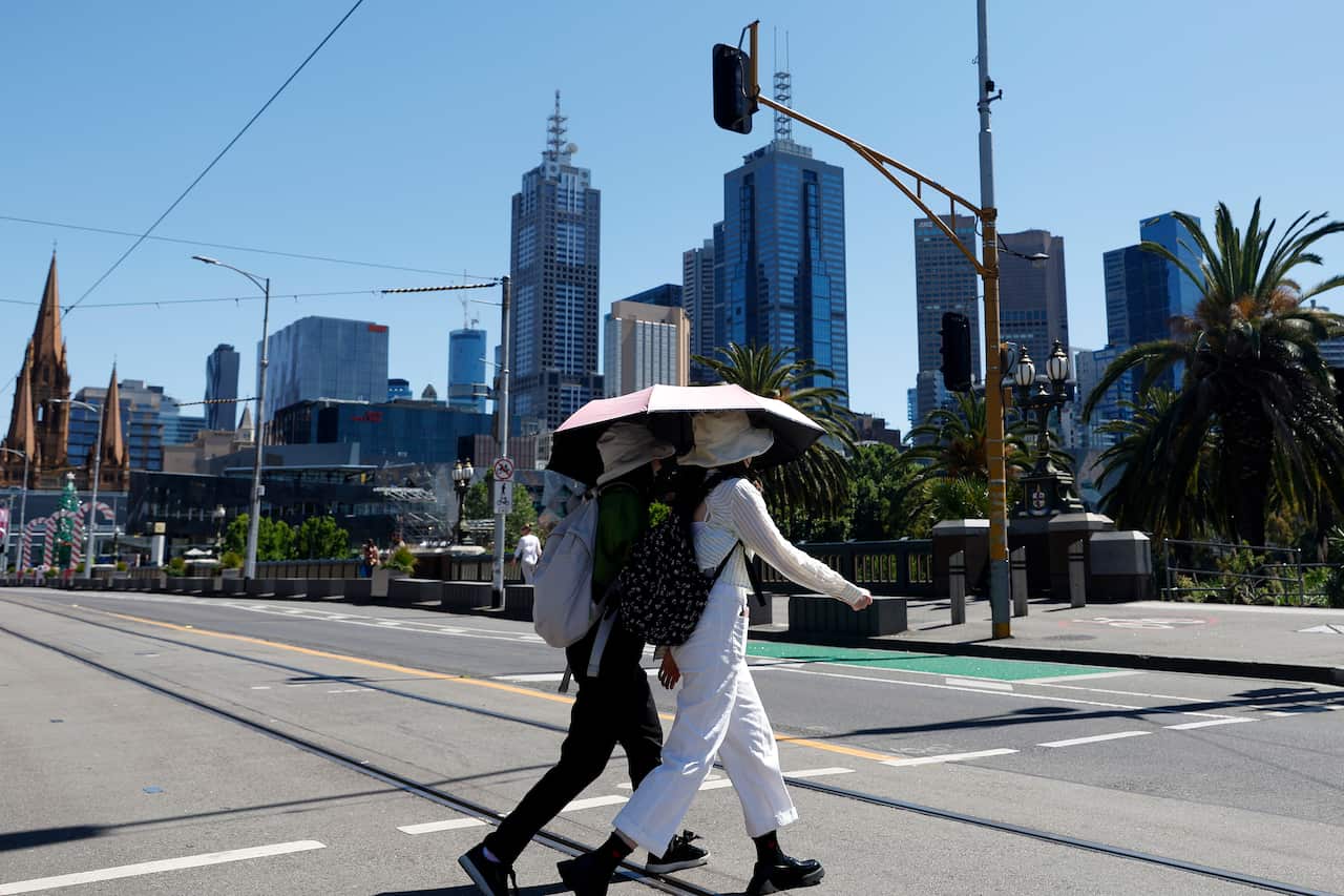 Two people wearing hats, backpacks, and long sleeve pants and shirts walk across the street while holding a sun umbrella, crossing the street. City buildings can be seen behind them. 