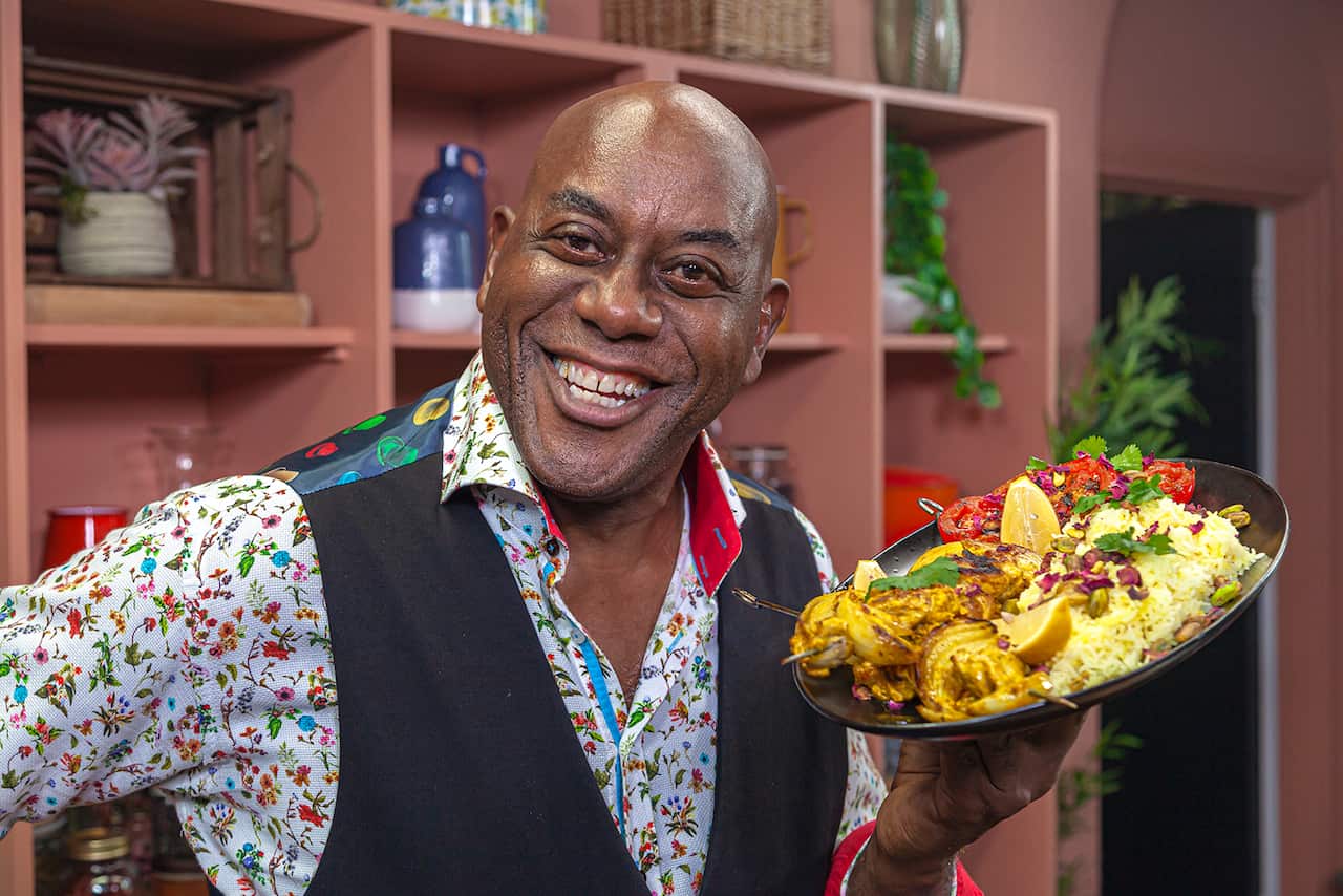 Man in white shirt with a floral pattern and dark waist coat smiles while holding a large oval plate with a chicken and rice dish. 