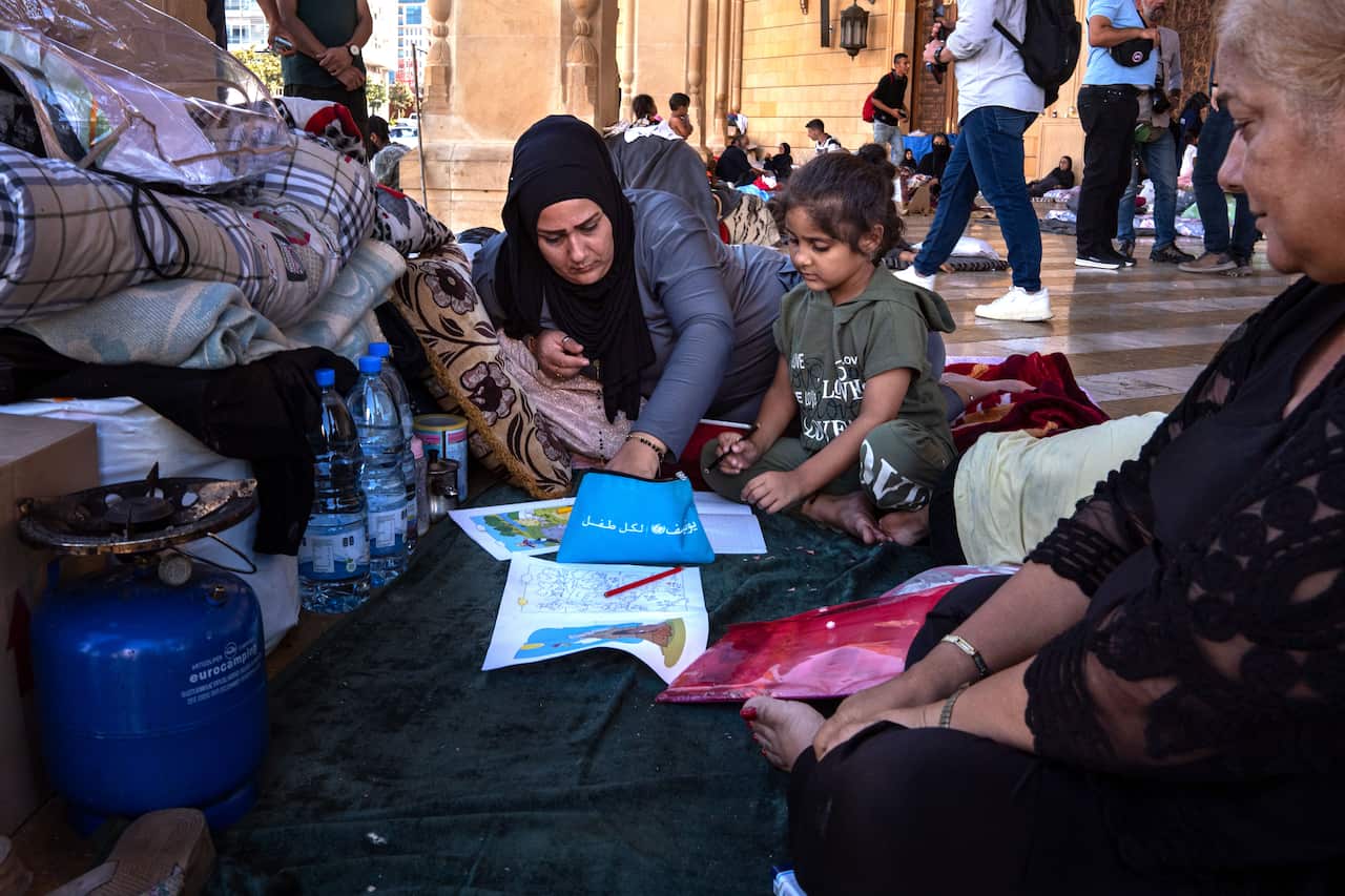 A woman wearing a headscarf reaches into a pencil case with Arabic writing on it. A young girl is sitting next to her and several sheets of paper with drawings on them sit in front of them. Several bottles of water and a small portable gas stove are next to them.