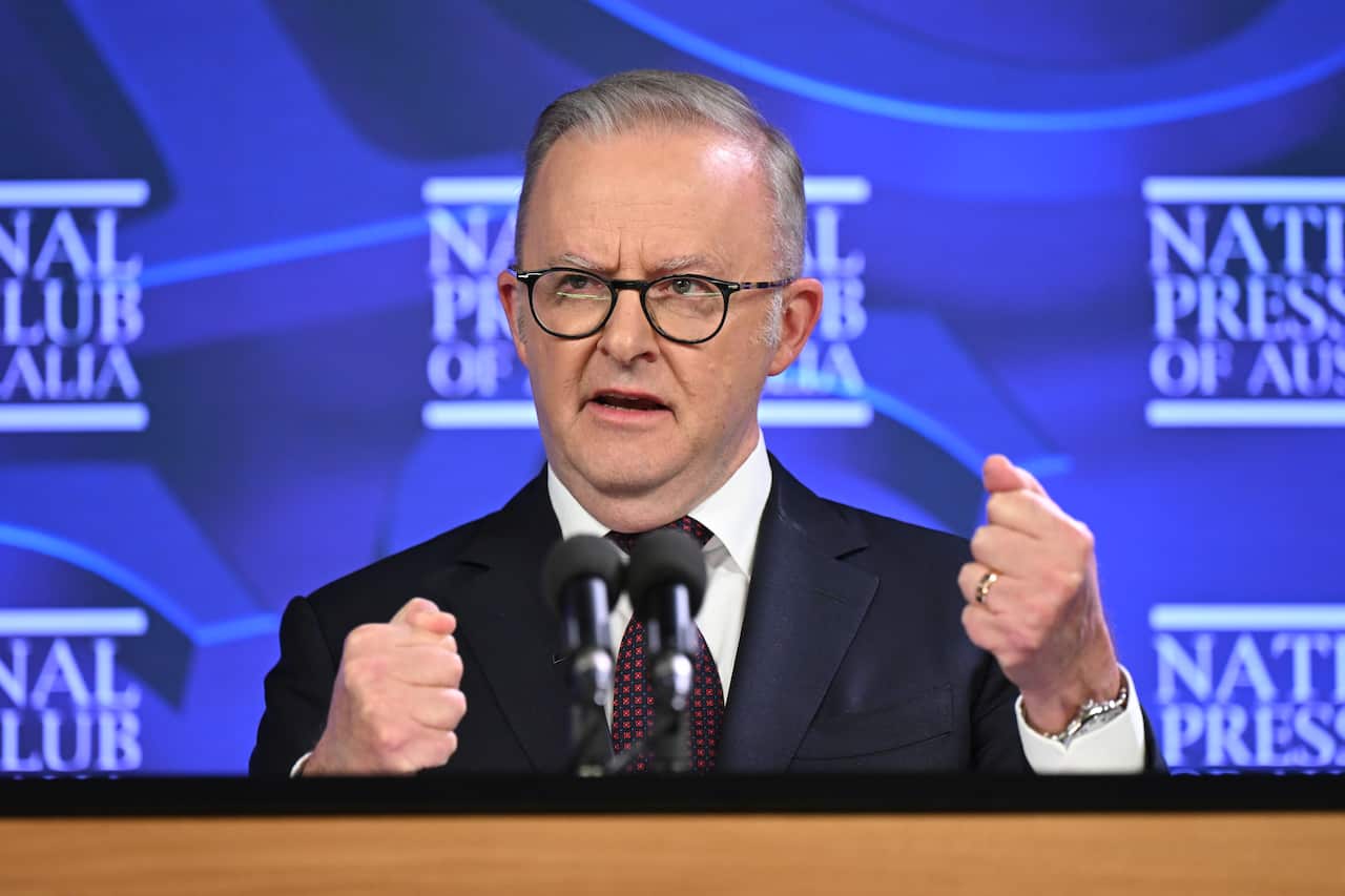 Prime Minister Anthony Albanese delivers an address to the National Press Club in Canberra. He stands behind a brown lectern with the blue backdrop of the press club behind him. He wears a dark suit with a red patterned tie and rounded glasses on his face.