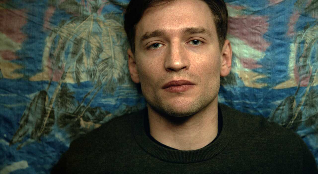 The head and shoulders of a young man are seen from overlead, lying on a patterned fabric. 