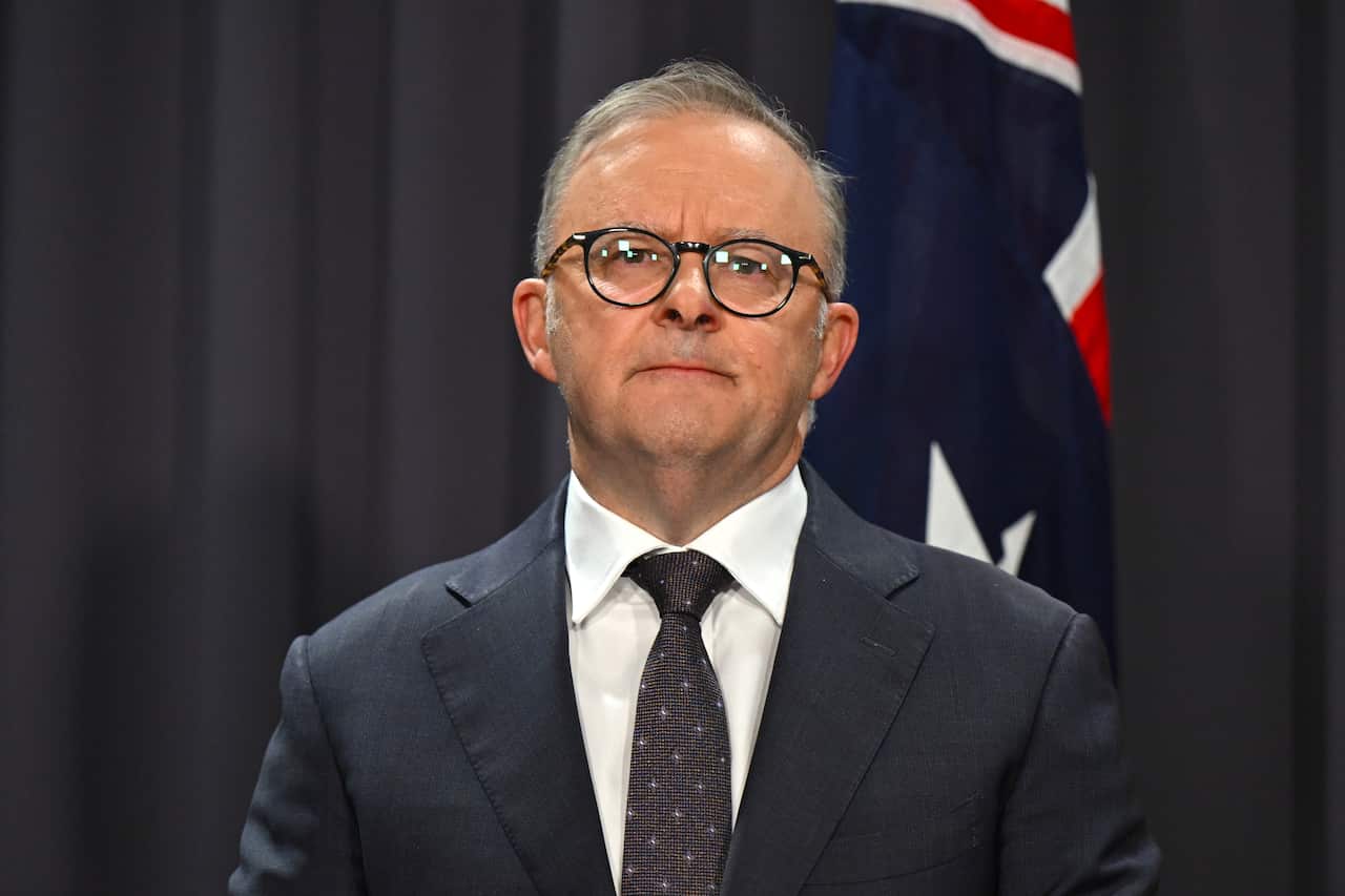 A man in a black suit and white shirt stands in front of an Australian flag. 