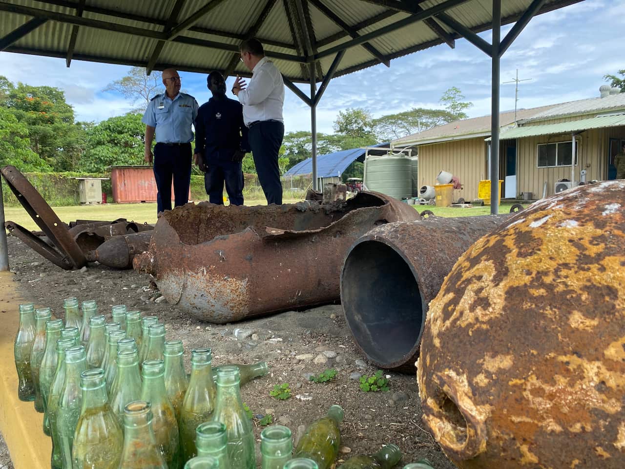 Three men talking. In front of them are rusted military shells.