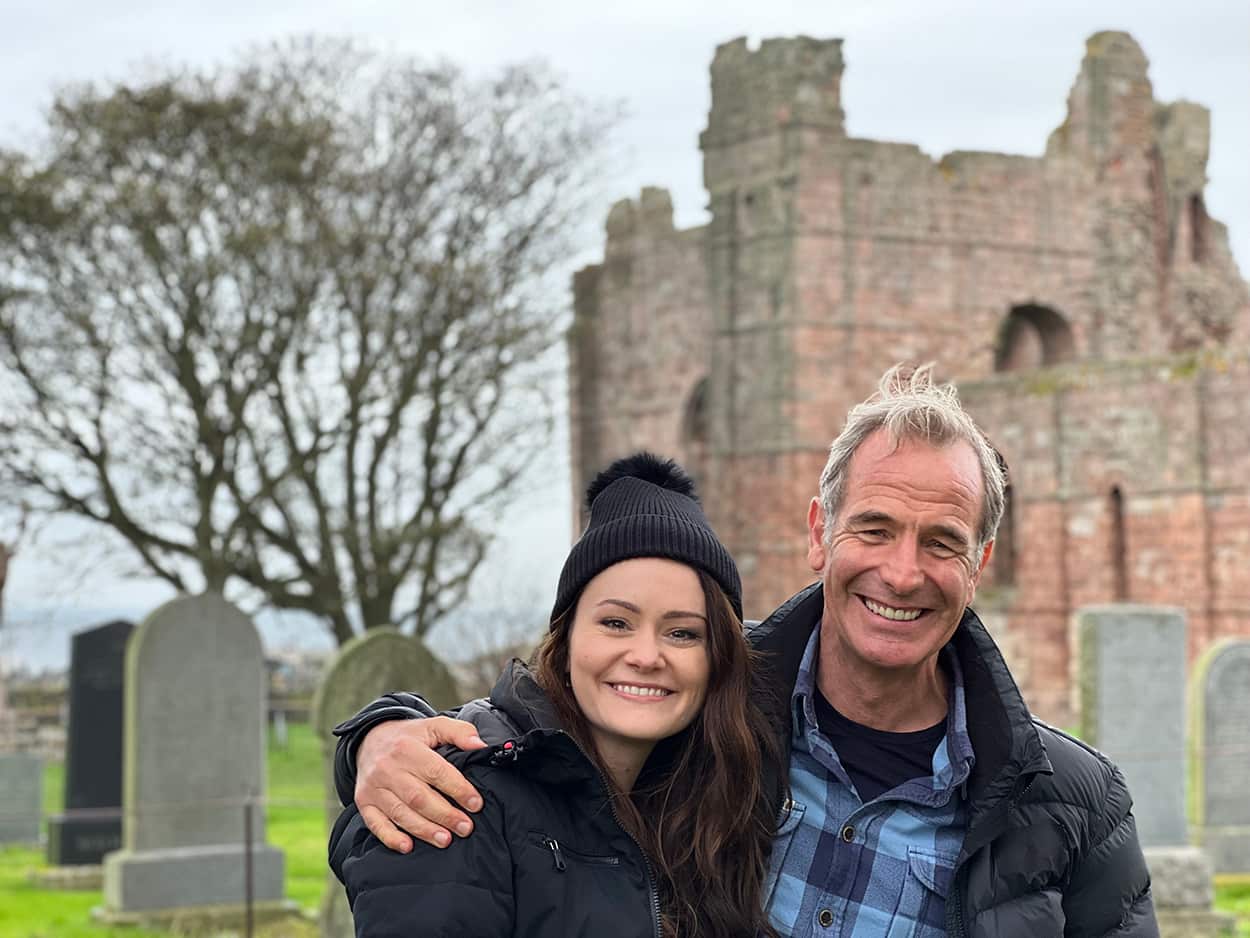 A man stands with his arm around a woman in the grounds of a ruined castle and cemetery in rural England.jpeg