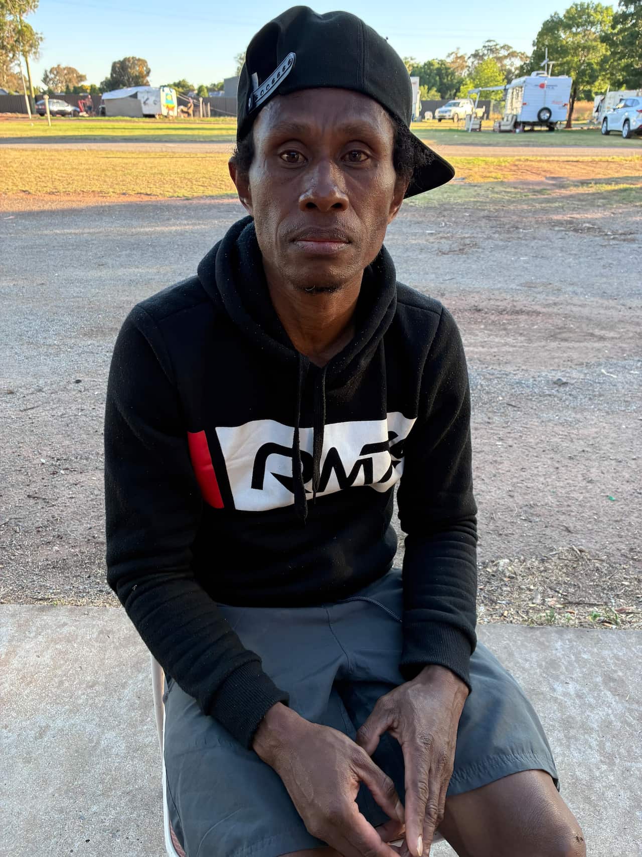 A brown-skinned man wearing a black hoodie and cap sits in a park.