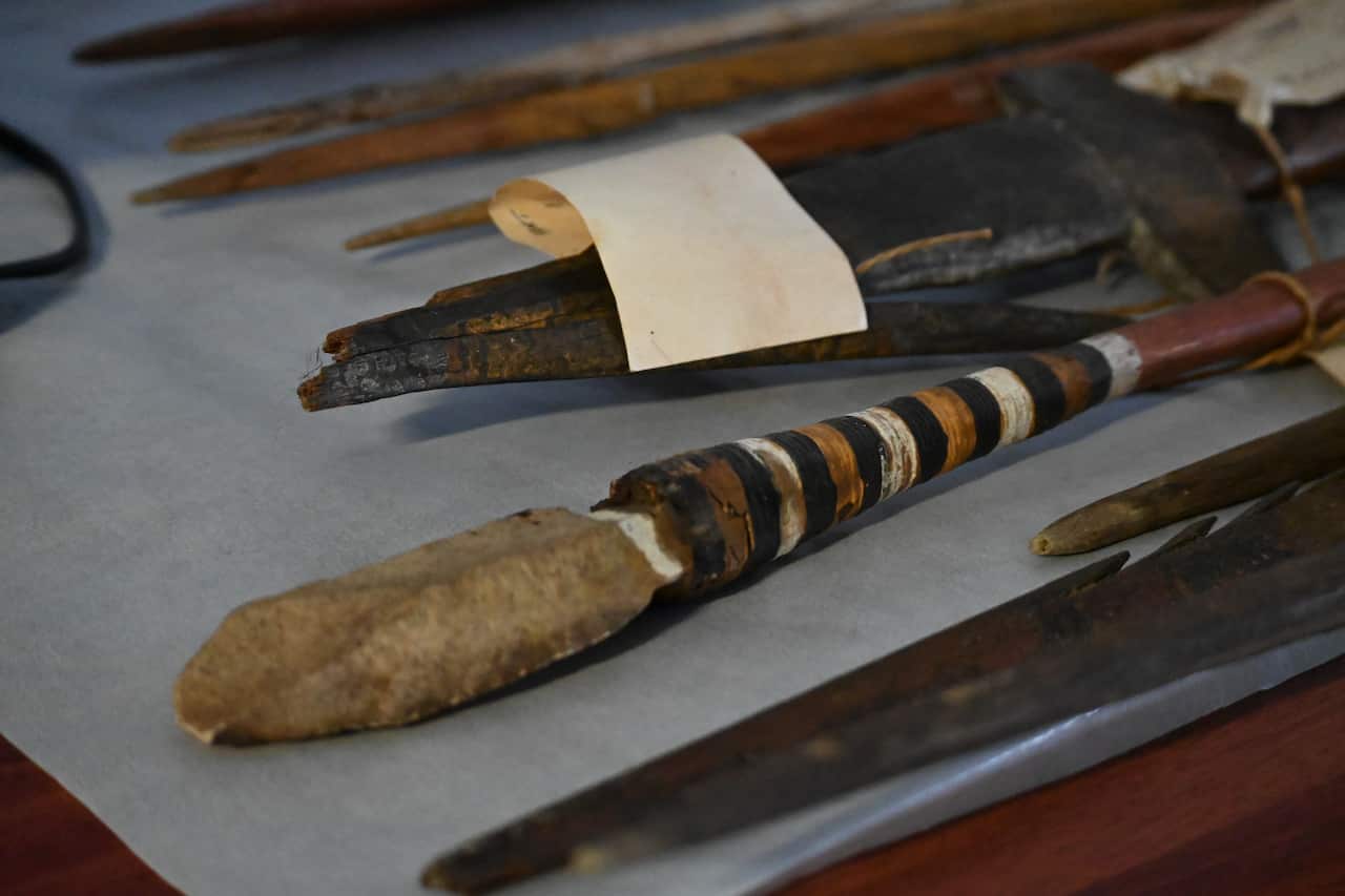 A collection of spears are lined up on a bench. They are all different. Some are carved timber, others are made with a stone head.