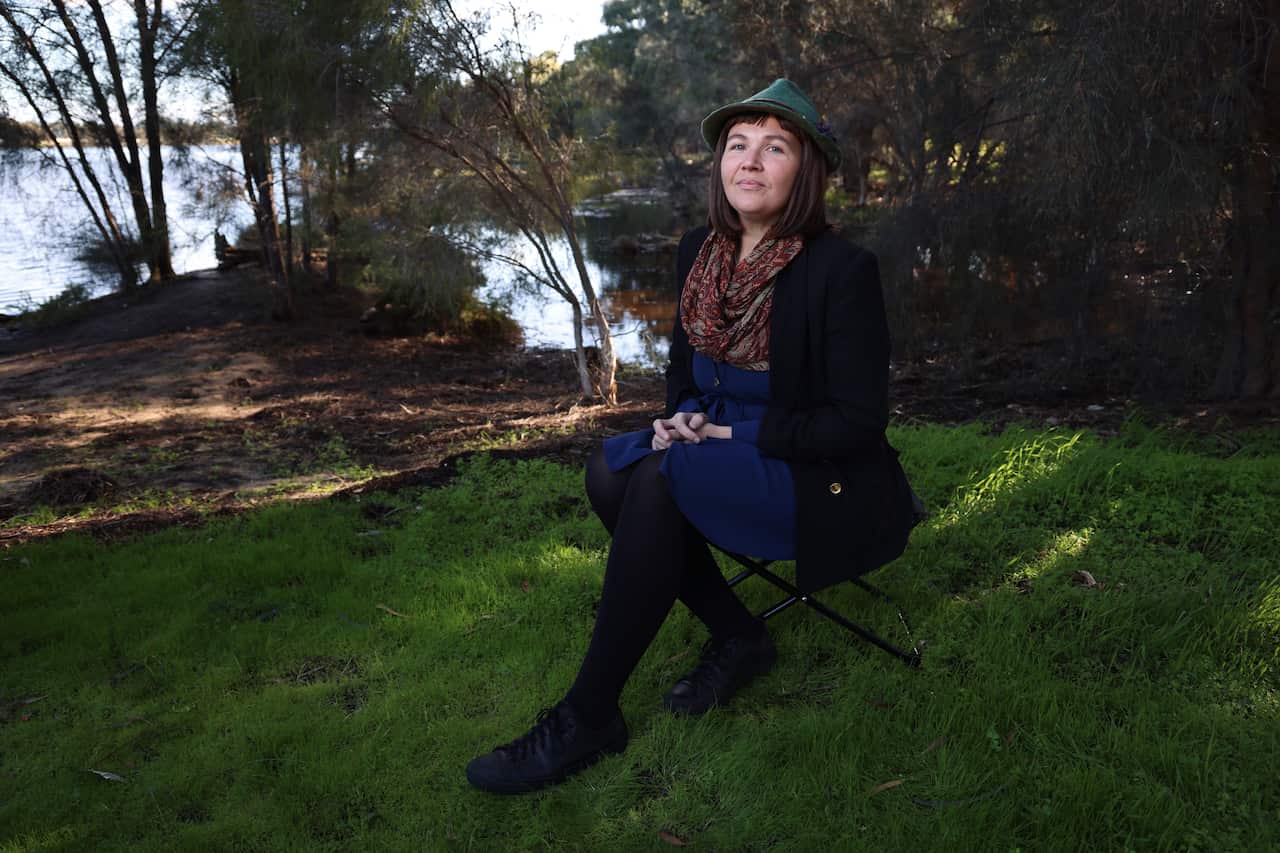 A middle-aged woman poses for a photo, sitting in a garden next to a water body.