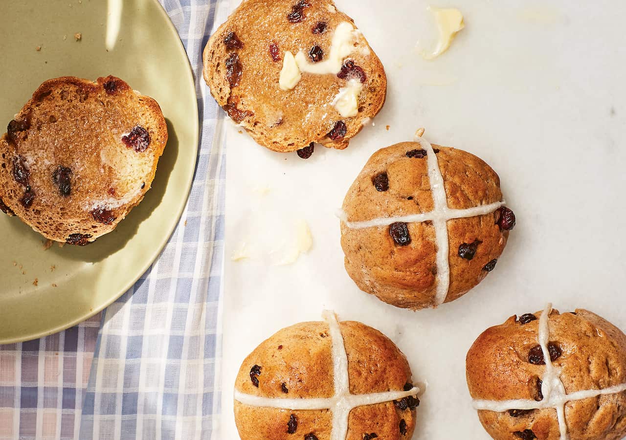 Seen from overhead, three hot cross buns sit on a white board. A fourth has been toasted and split open, with half on the board and the other half on a green plate that sits alongside. The bottom of the bun has melting butter on it. 
