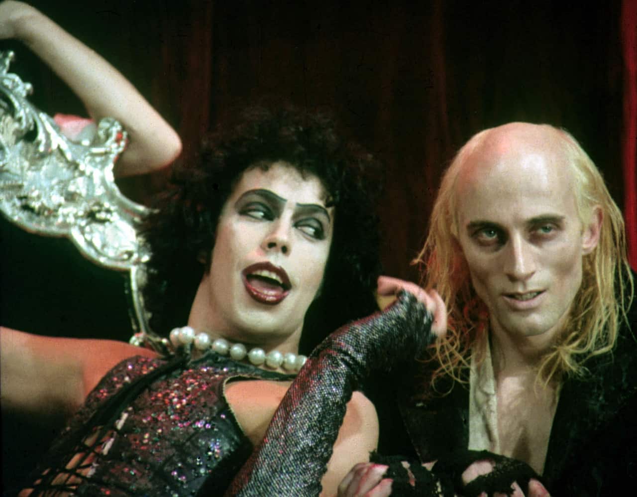 Two performers in elaborate stage costumes seated together against a dark backdrop, with one wearing theatrical makeup, fishnet sleeves and a pearl necklace.