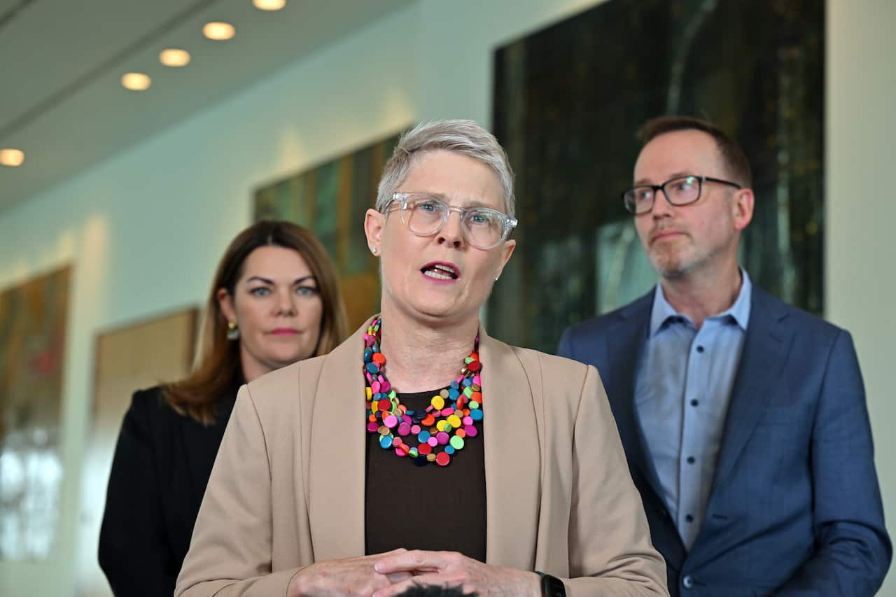 A woman with short hair in a tan jacket and a colourful necklace in front of two people in the background.