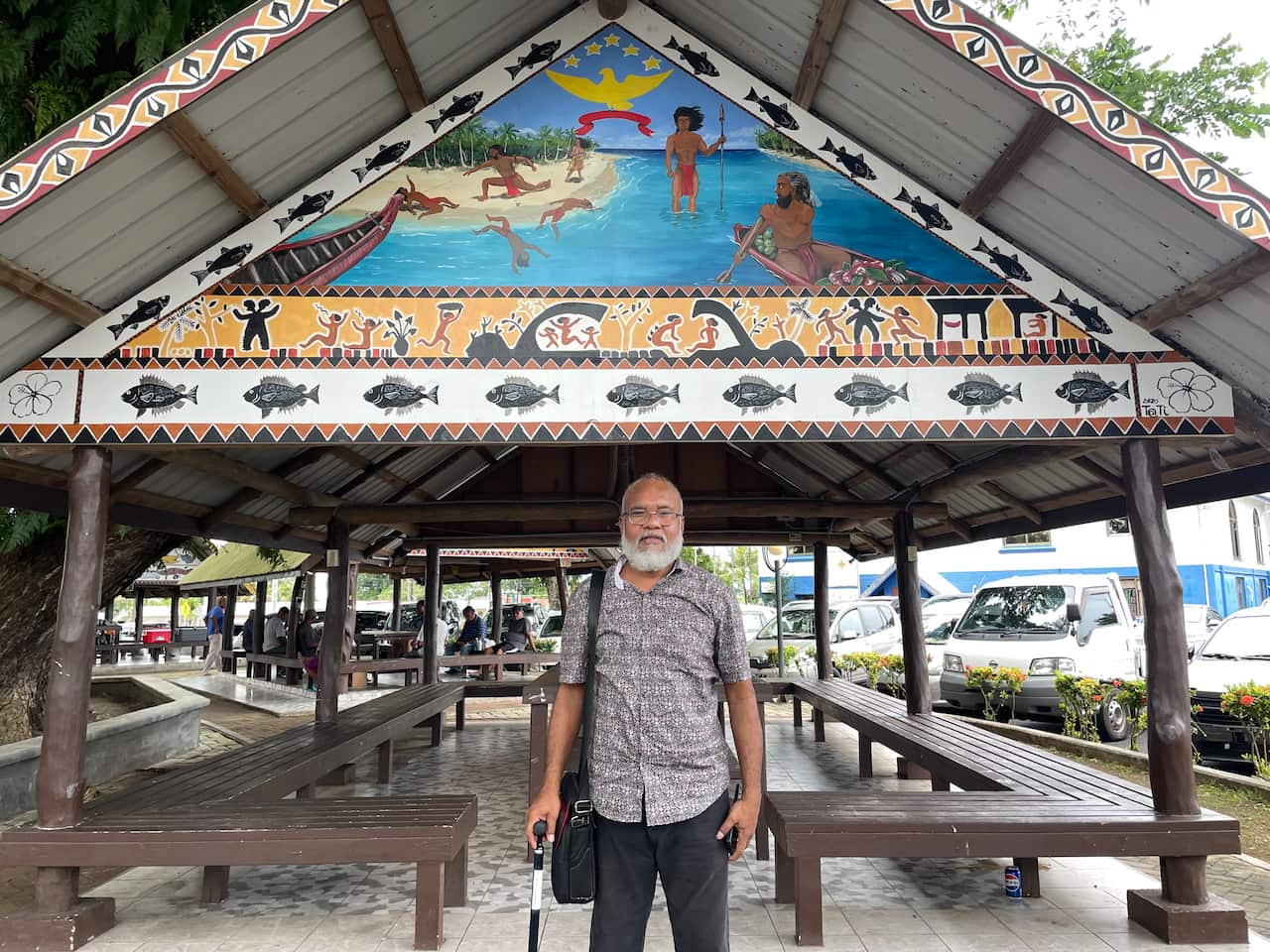 A man with a grey beard and a walking cane stands under an open-air pavilion adorned with intricate indigenous paintings and fish motifs along its gable.