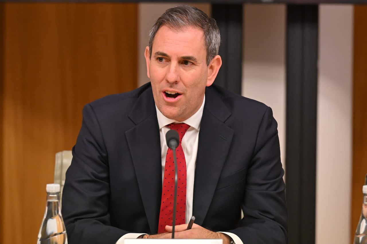 A man in a navy suit jacket and red tie speaks into a microphone. A bottle of water sits in the foreground.