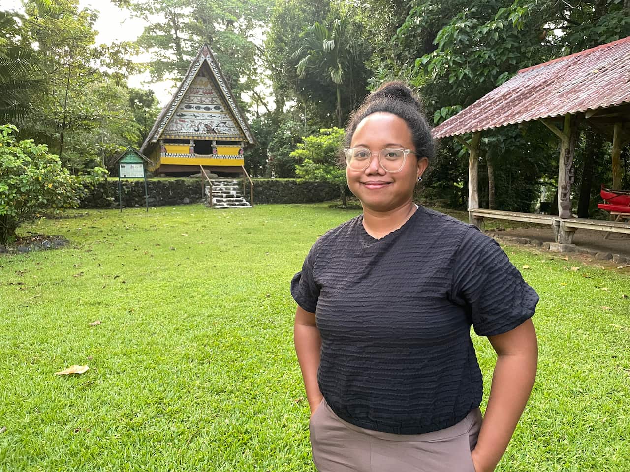 A smiling woman wearing glasses and a black top stands on a grassy lawn in front of a traditionally decorated wooden structure nestled among lush foliage.