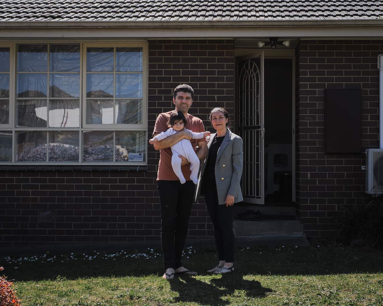 A man, woman and baby stand outside a brick house, facing the camera. The man holds the baby in his arms while the woman stands next to him.