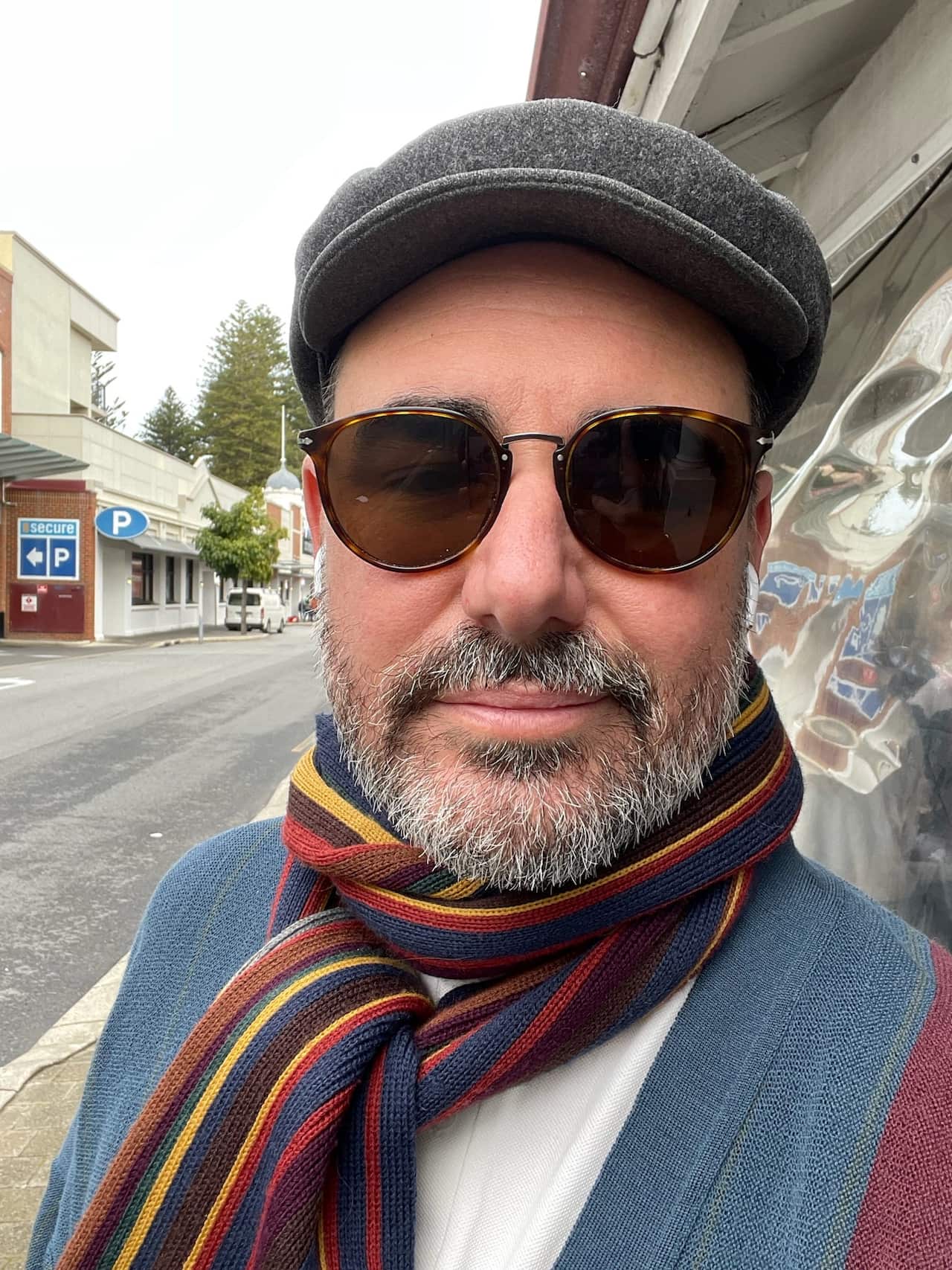 A man with a greying beard wearing a flat cap, sunglasses and multicoloured scarf stands in front of a painted mural on a street