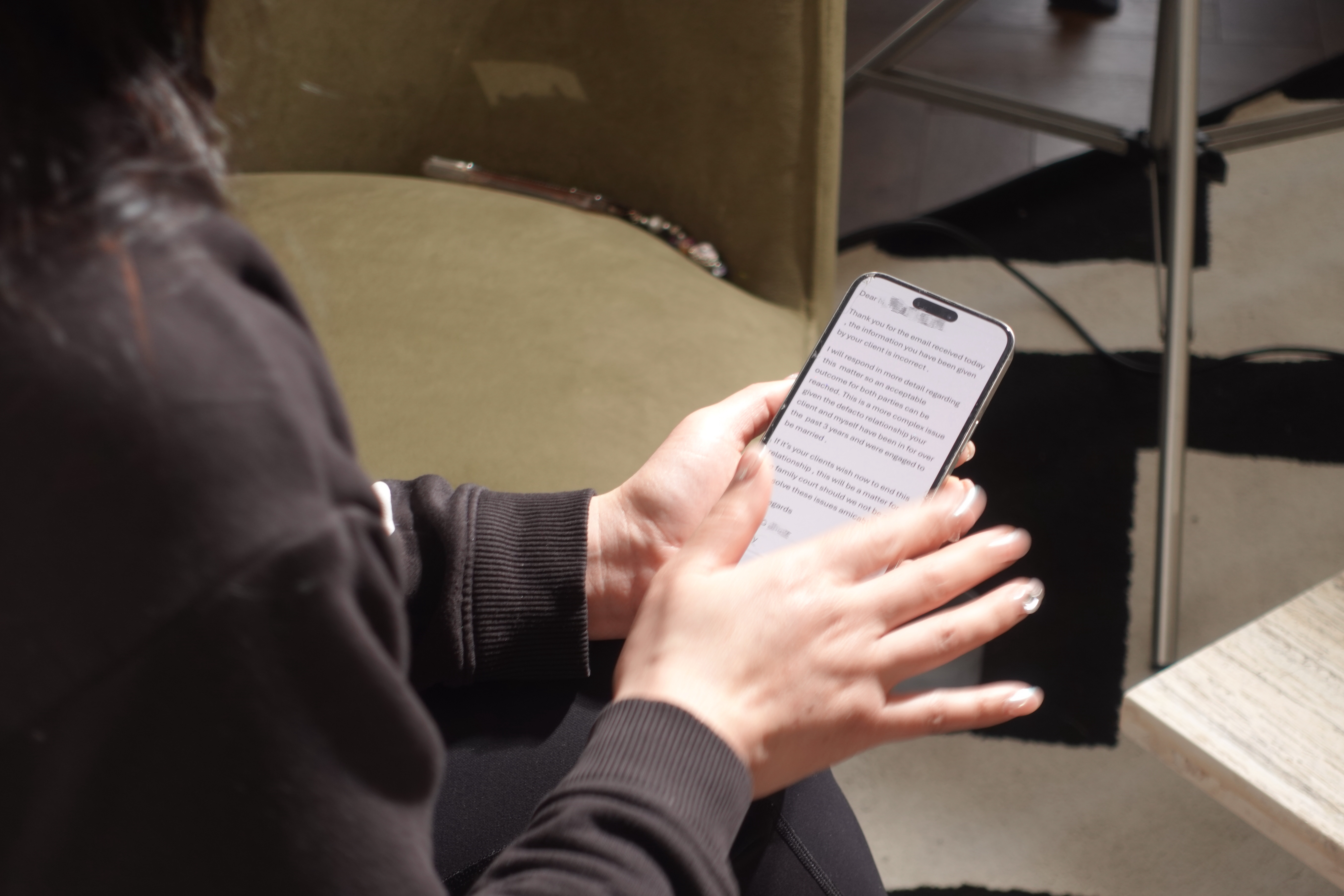 A person wearing a black sweatshirt and dark pants holds a smartphone and is reading an email displayed on the screen, with a blurred background of a moss-green chair and rug.