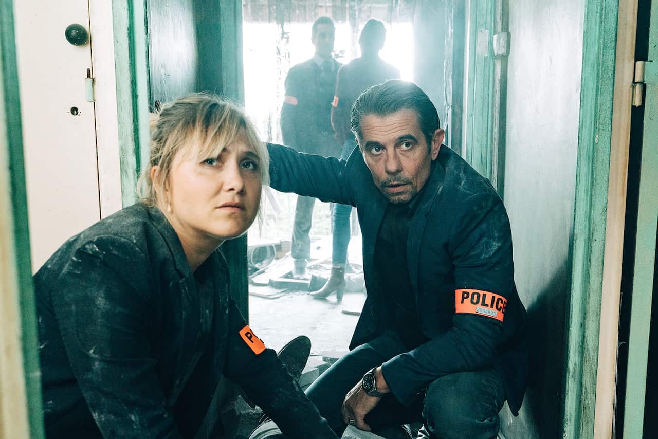 A woman and a mad crouch in a doorway. Both have orange armbands with the words 'POLICE' in them. Two more people, also with orange armbands, can be seen behind them. 