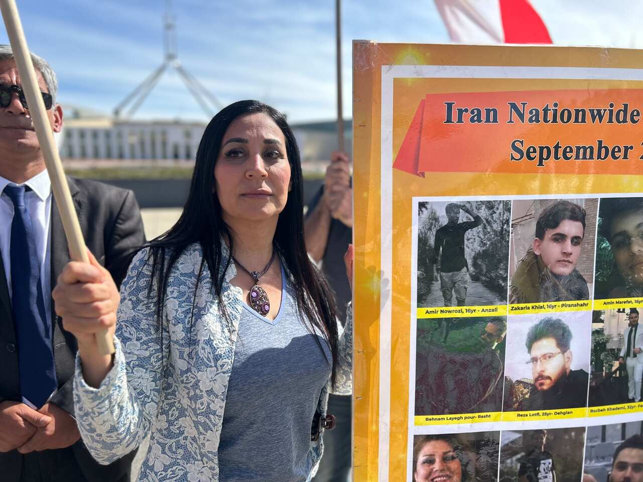 A woman at a rally standing next to a poster featuring people's faces. Parliament House in Canberra is in the background