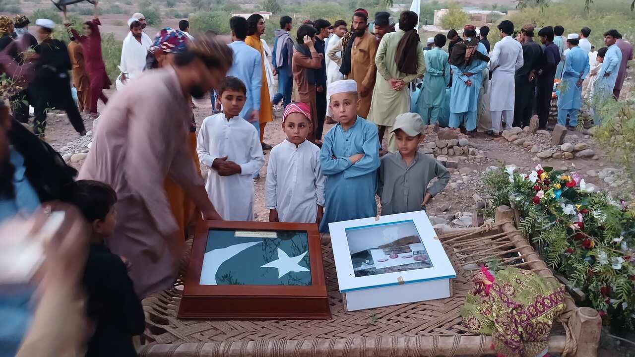 Four children are standing next to a charpai laden with two wooden boxes. A man is crouched on the string-bed trying to open one of the boxes. There's a crowd of men standing behind them.