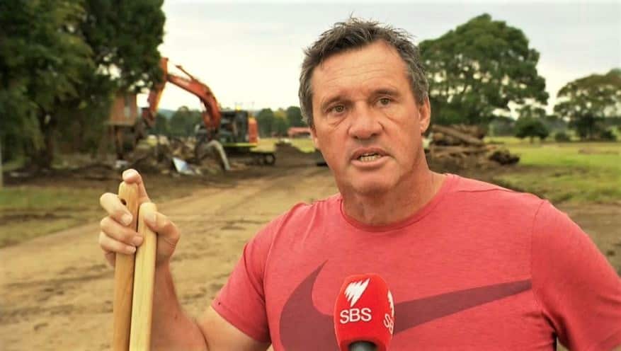 A man in a red shirt holds some tools in front of a digger, speaking into an SBS microphone.