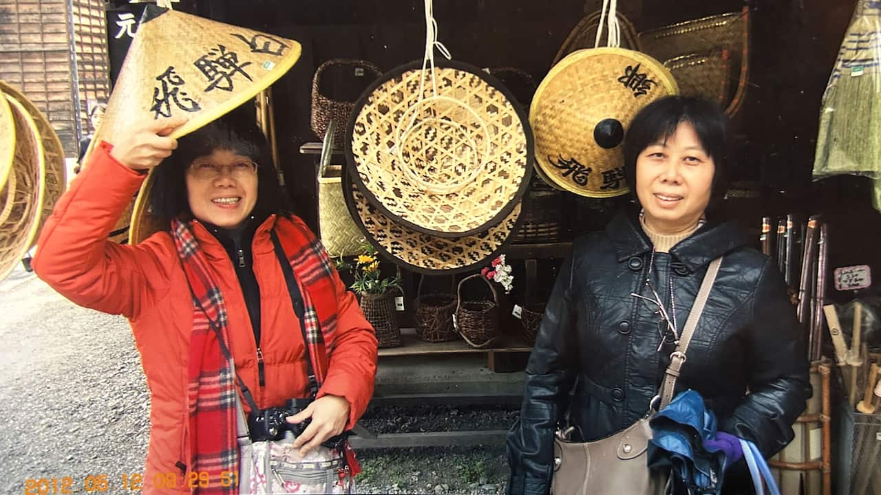 Two middle-aged Asian women wearing red and black puffer jackets and cross-body bags are posing for a photo with traditional conical straw hats with Chinese text on them