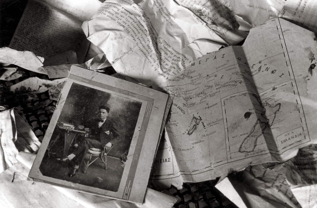 A black and white, high-angle photograph shows a scattered collection of historical artifacts, including a portrait of a seated man in a suit, a hand-drawn map with Greek annotations, and various crumpled documents.