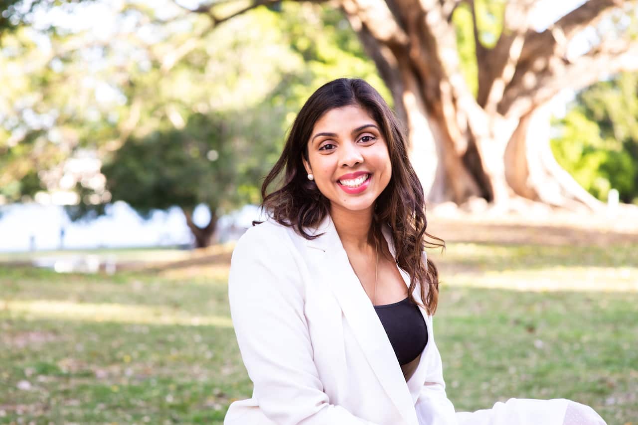 A woman in a white suit and long dark hair sits outside on the grass and smiles.