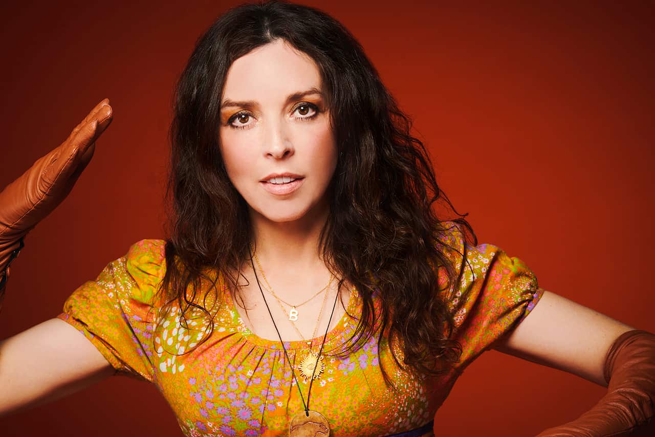 A woman in a bright patterned orange dress and long brown gloves poses dramatically, arms at angles.