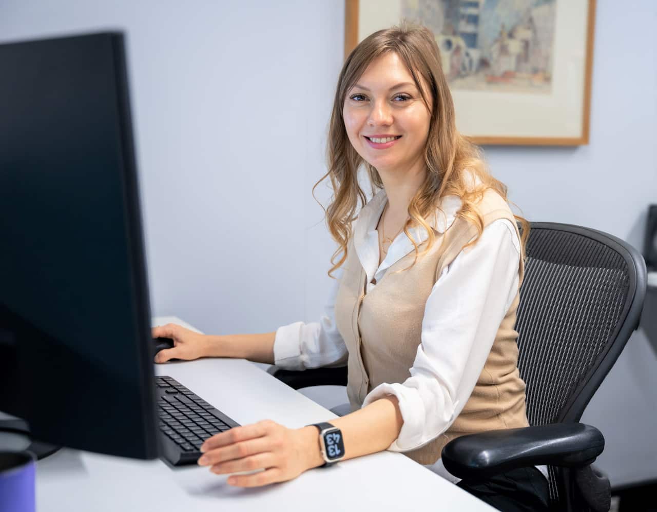 A woman with long brown hair wearing a beige vest sits at a dsk.