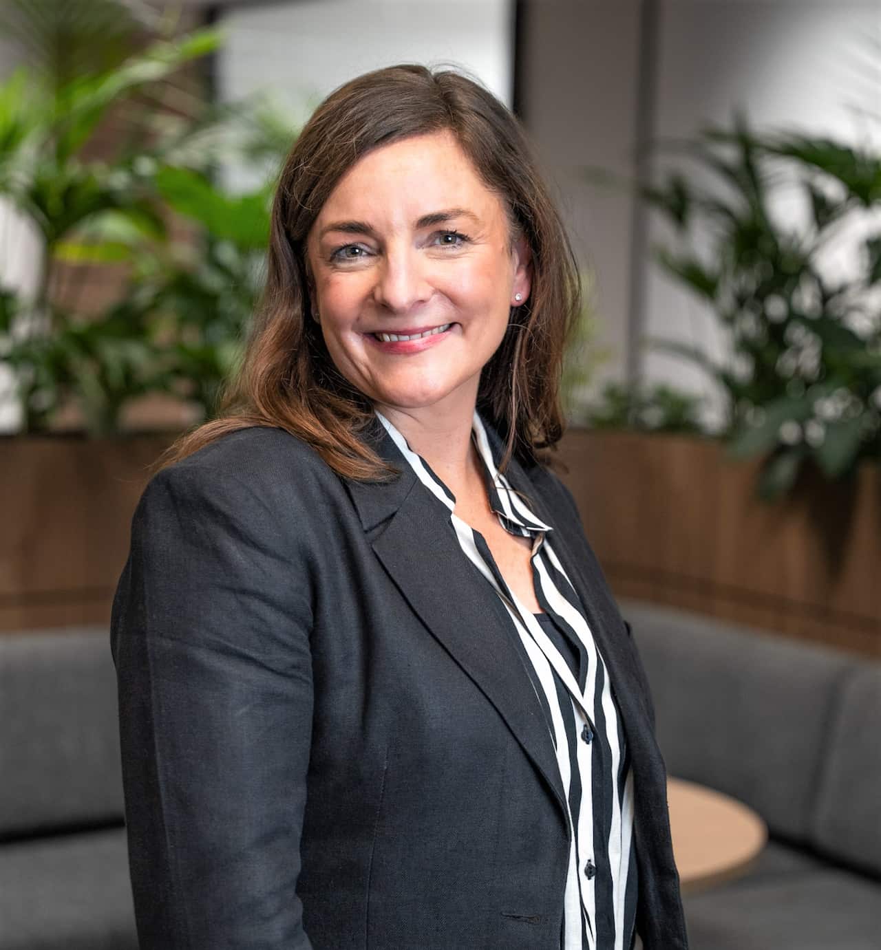 A woman in a blue blazer stands in an office facing camera.