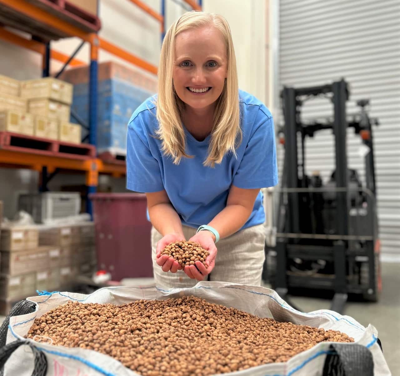 A woman in a blue t-shirt holds a handful of small, brown nut-like items above a large sack full of them.