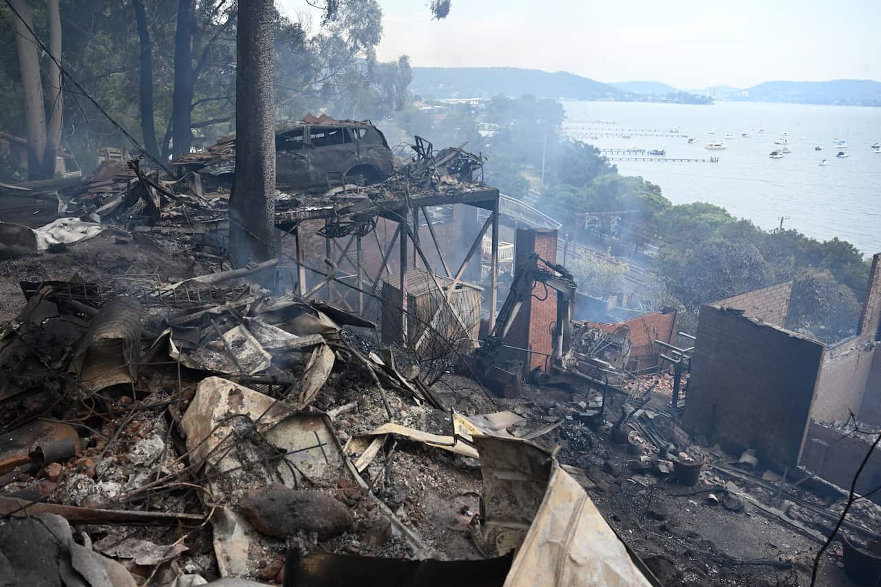 The wreckage of homes and a vehicle after a bushfire, with a bay visible in the background.