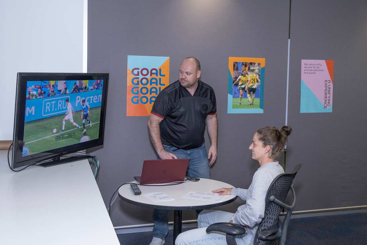 Alan March is wearing a black football jersey and standing next to a television screen showing a football match. A woman is sitting at a round table and also watching the screen.