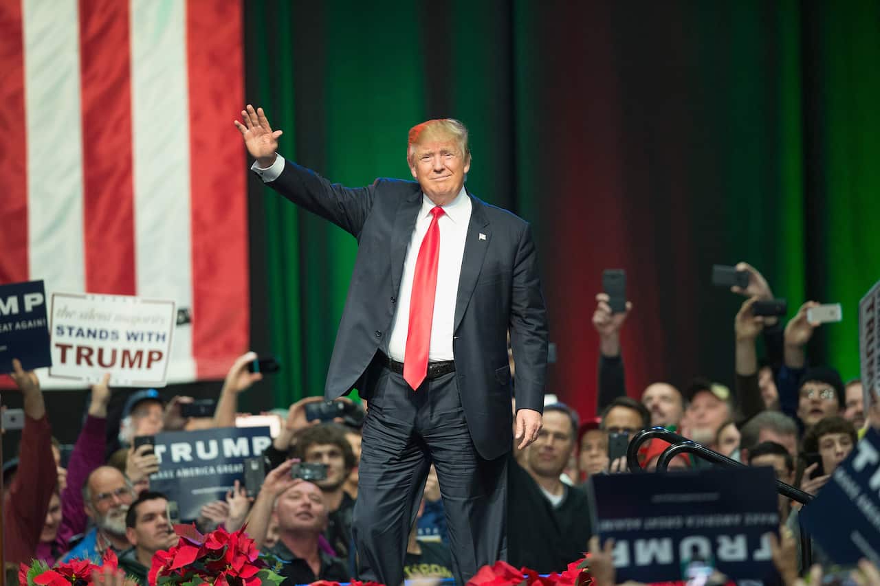 Donald Trump stands on stage, raising his right hand to wave at the crowd during a campaign event. In the background, attendees are capturing the moment with their phone cameras, holding signs that read Stand with Trump and others.