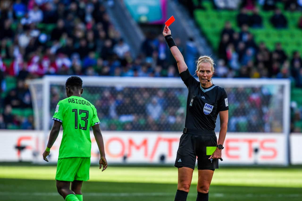 A woman in a black referee uniform holds up a red card to a woman in a fluorescent green football uniform.