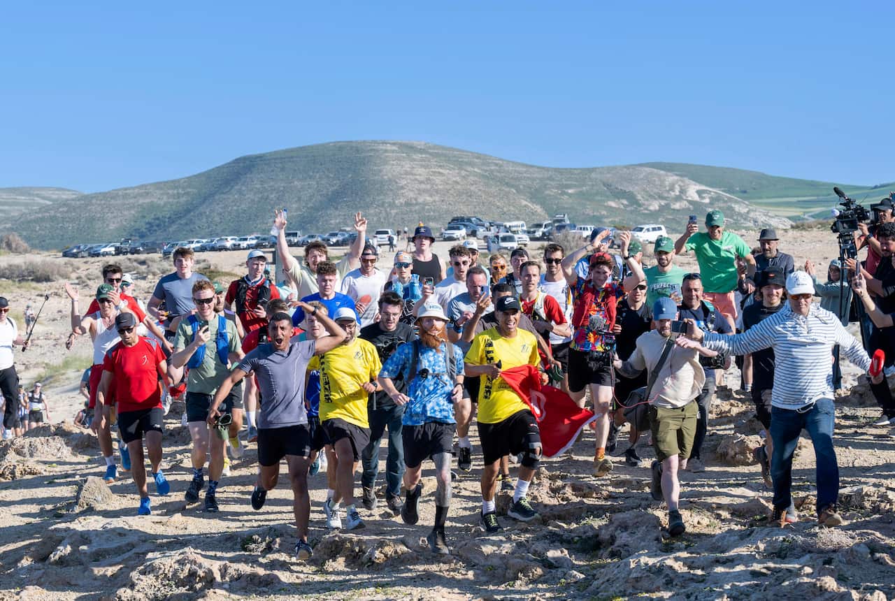 Russ Cook with a group of supporters in running across sandy ground in Tunisia. There is a large green hill in the distance.