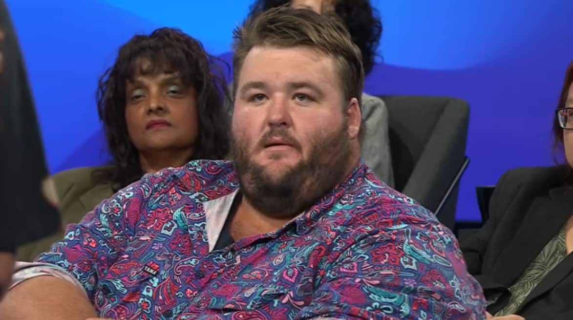 a man in a pink and purple patterned top in a studio audience with other members in the background
