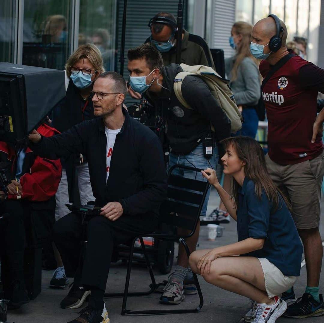 A group of crew members and actors, several wearing face masks, gather around a camera monitor on a film set to review a shot.