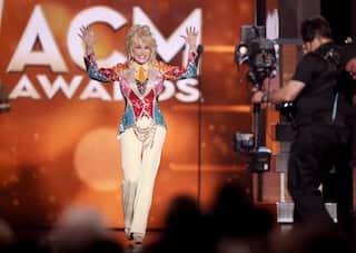 Dolly Parton in a brightly coloured sequined outfit walks onto an awards-show stage with both hands raised, as a television camera operator films nearby.