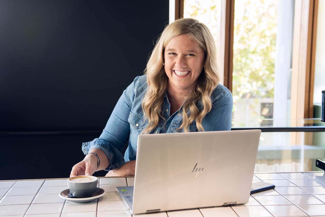 A smiling woman sitting at a bench with a coffee and laptop in front of her.