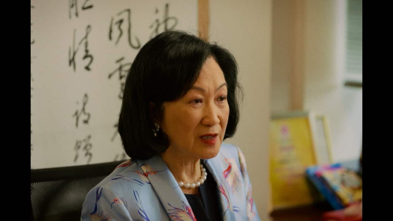 An older Asian woman with dark hair speaking during an interview while wearing a light blue floral suit with a pearl necklace