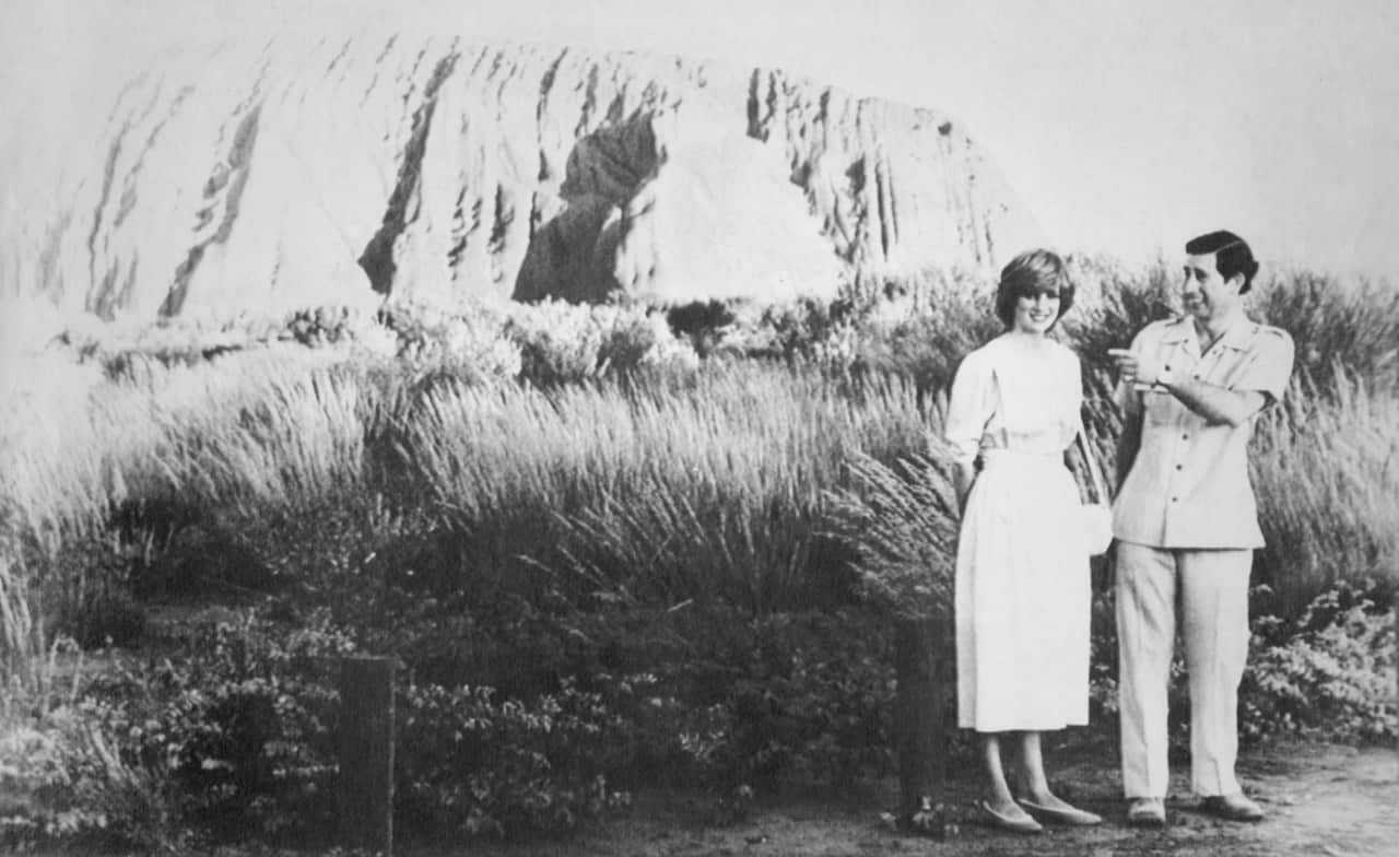 A black-and-white photograph shows Princess Diana and Prince Charles standing in a grassy field with the massive rock formation of Uluru in the background.