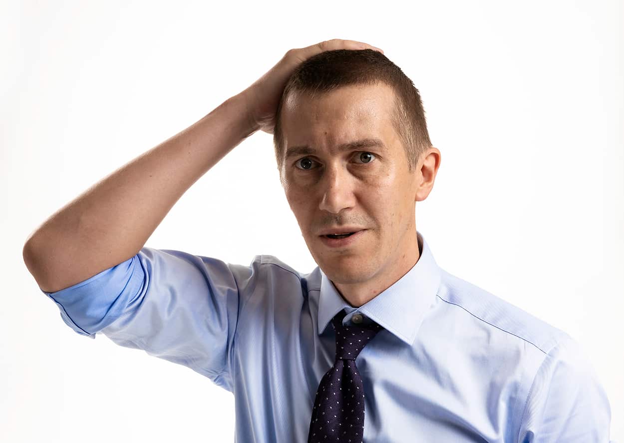 A man with a bewildered expression stands with his hand on his head.
