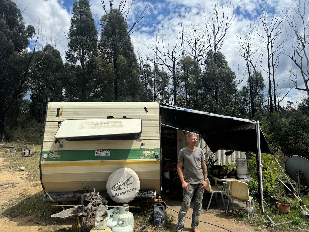 A man is standing in front of a caravan parked in an area surrounded by trees.