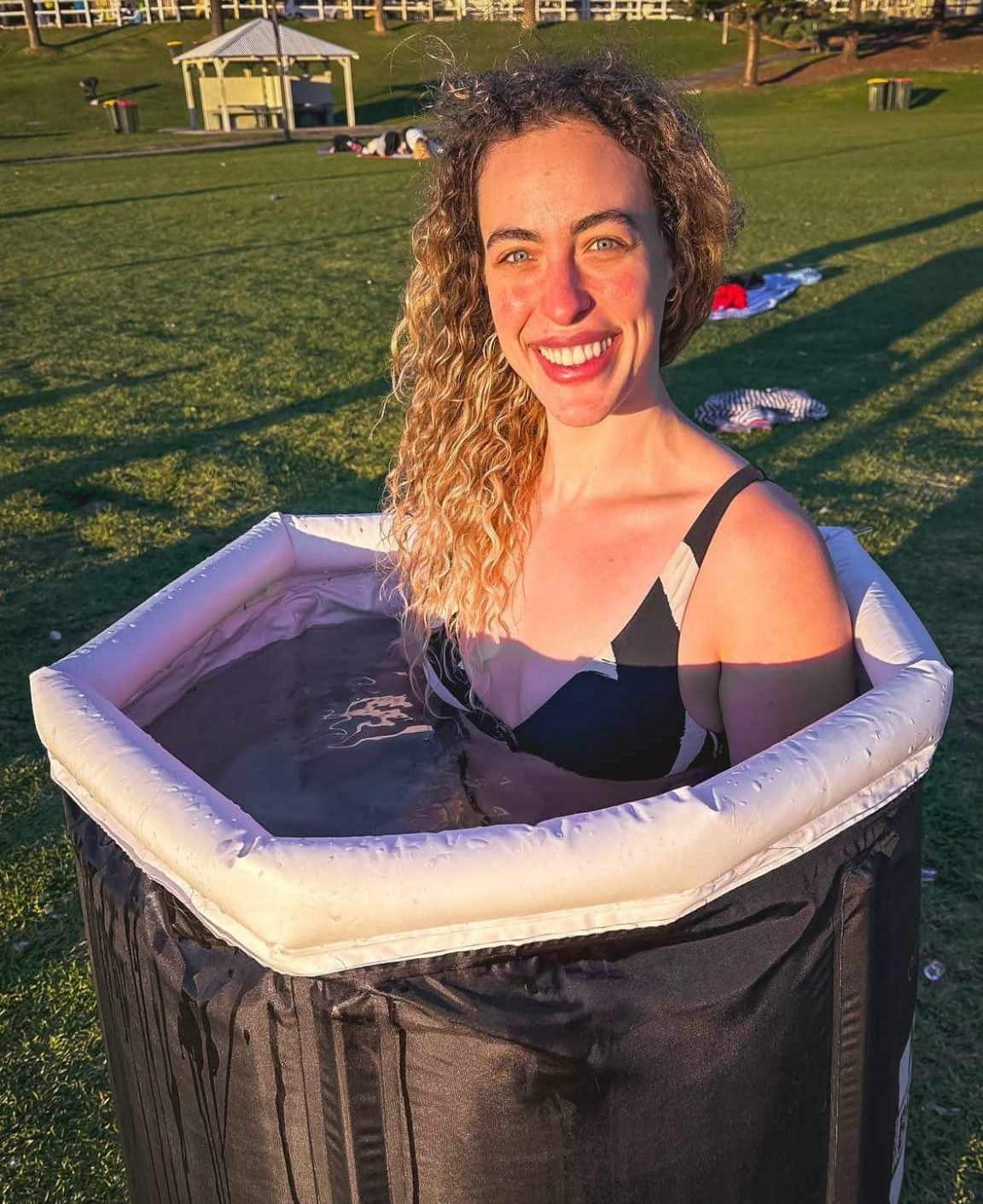A woman in a bikini sits in a tub of ice and smiles.