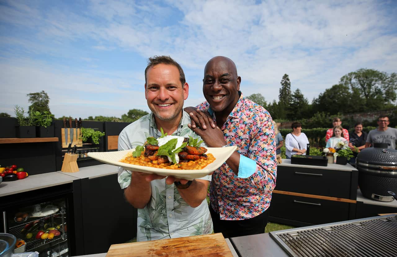 Ainsley Harriot and Tristan Welsh stand in an outdoor kitchen, smiling. 