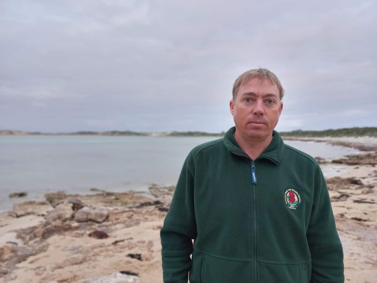 Aaron Smith, a ranger standing on the coastline of South Australia, wearing a green jumper, he looks serious.