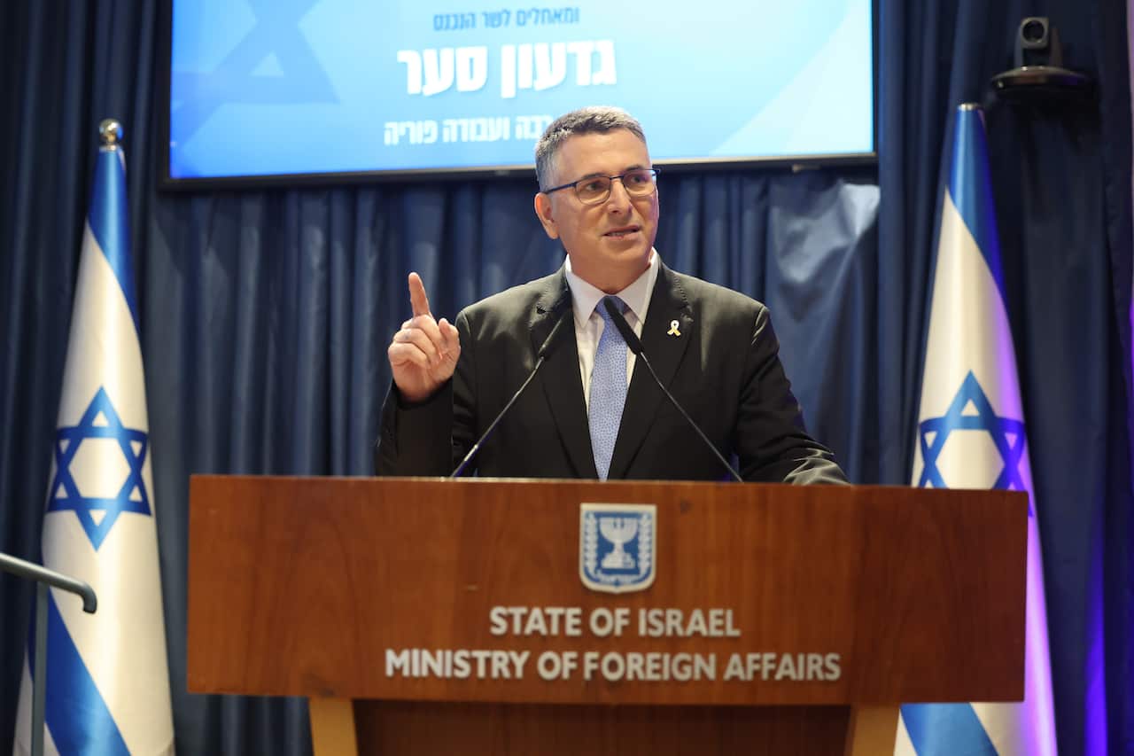 A man in a dark suit and light blue tie speaks from a podium.