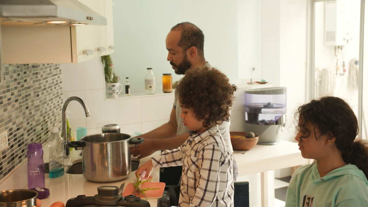 A man at the kitchen cooking a meal with his two children, a boy in a checkered shirt and a girl in a mint-colored hoodie