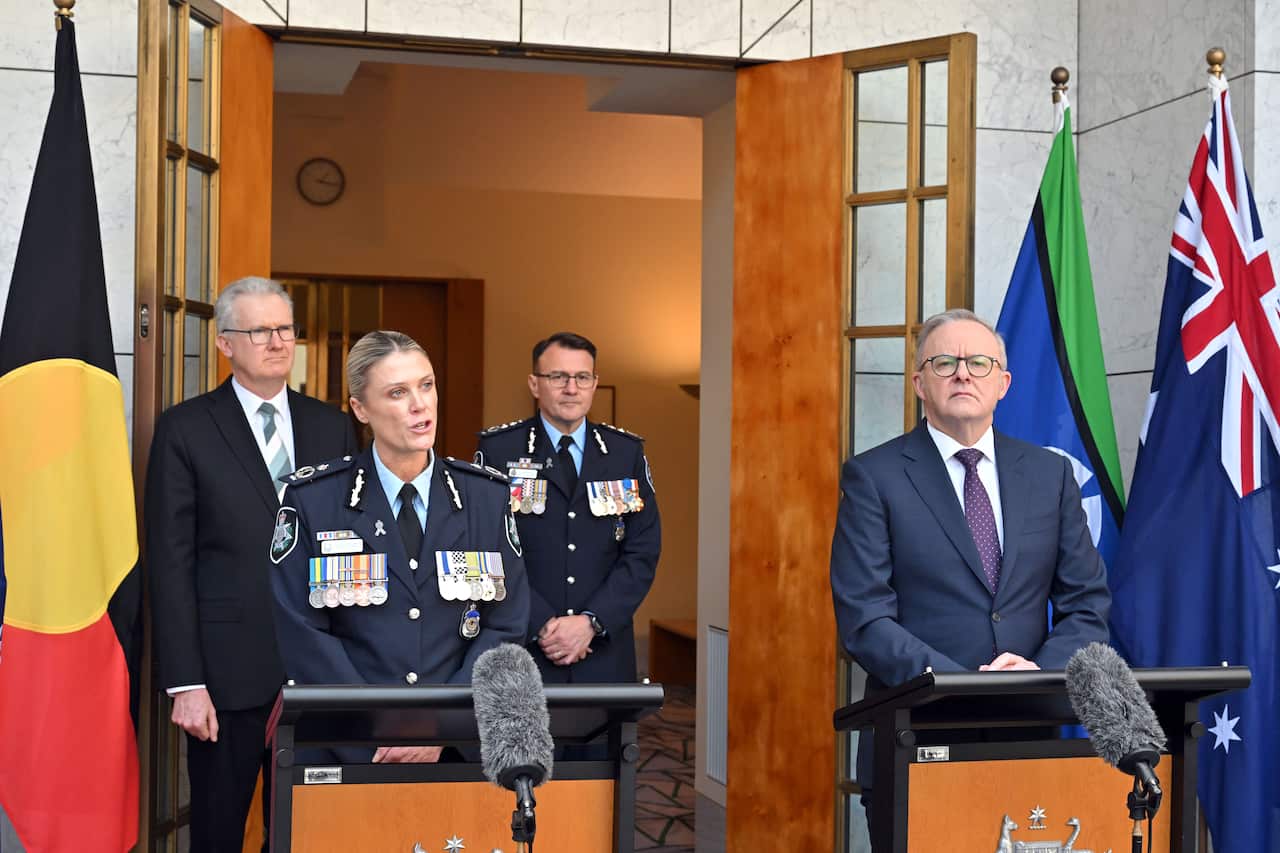 Anthony Albanese and Krissy Barrett stand at side-by-side podiums in front of a set of open doors, with Australian, Torres Strait Islander and Aboriginal flags, as well as two other people, behind them. 